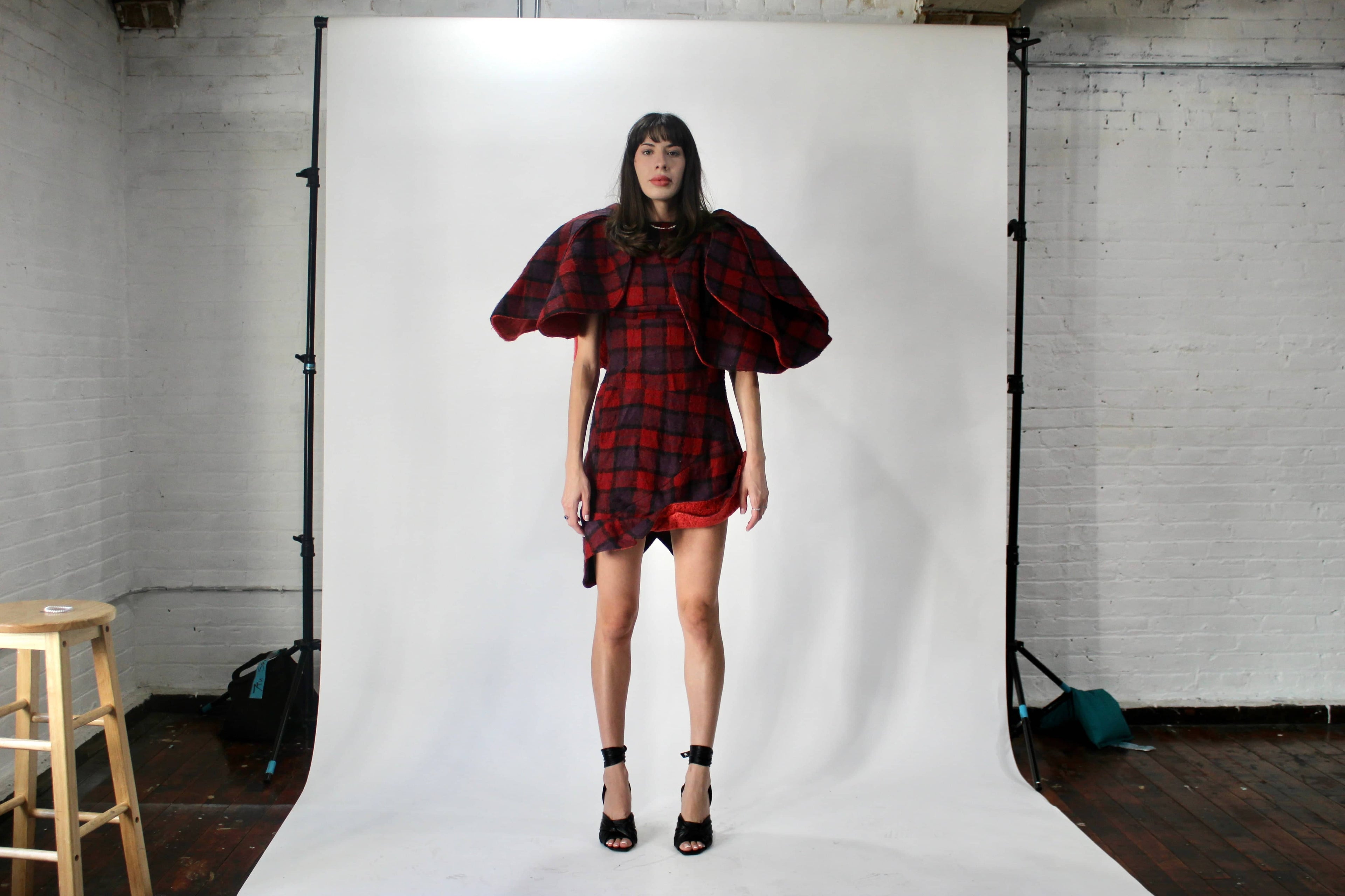 Model in a behind-the-scenes fitting wearing a Corii Burns plaid wool dress with sculptural flower shoulder sleeve, part of the MHII Collection for NYFW prep in NYC studio.