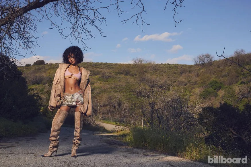 South African singer and songwriter Tyla Laura Seethal wearing the Corii Burns Sante Mentale SS24 Skirt for Billboard Magazine while standing on concrete back country road while wearing large beige overcoat and high heeled boots; A striking woman in a chic bikini top and fashionable boots stands poised on a rustic dirt road, radiating bold elegance.